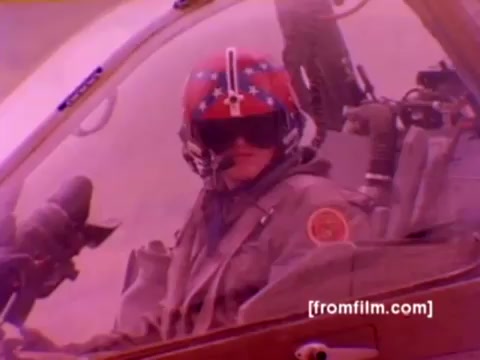 The still from the 'USMC Aviation Ordnance enlistment film' features a pilot in the cockpit of an AH-1 Super Cobra helicopter, characterized by a distinctive helmet adorned with stars and stripes. The image likely captures a moment of preparation for flight, showcasing the advanced technology and weaponry associated with Marine Corps aviation. The background includes various aircraft like F-4 Phantoms, FA-18s, and Harriers, highlighting the ordnance option for enlistment. The overall tone is muted, reflecting the 16mm film's vintage quality, but color correction would enhance the vibrancy of the scene and aircraft details.