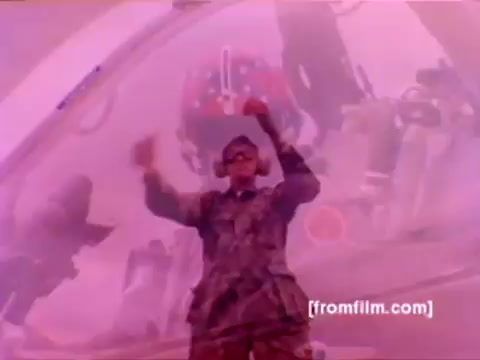 The still from the 'USMC Aviation Ordnance enlistment film' showcases a Marine in a uniform, focusing on ordnance operations. In the background, military aircraft like F-4 Phantoms, AH-1 SuperCobras, an FA-18, and a Harrier are represented, suggesting an active airfield environment. The Marine appears to be involved in the process of mounting missiles and bombs, indicating hands-on work with ordnance. The color correction would enhance the visibility and vibrancy of the military setting, improving the overall clarity of the scene.