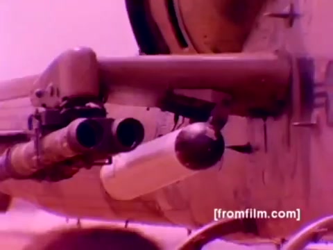 The still from the 'USMC Aviation Ordnance enlistment film' captures a close-up view of ordnance being mounted on a military helicopter, likely an AH-1 Super Cobra. The image showcases missile pods attached to the aircraft, along with a suspenseful aura conveyed by the film's color grading, which features a reddish tint. The focus on ordnance highlights the technical aspects of aviation support within the Marine Corps. This scene emphasizes the importance of the Ordnance Option in military enlistment, showcasing the equipment and readiness of Marine aviation units.