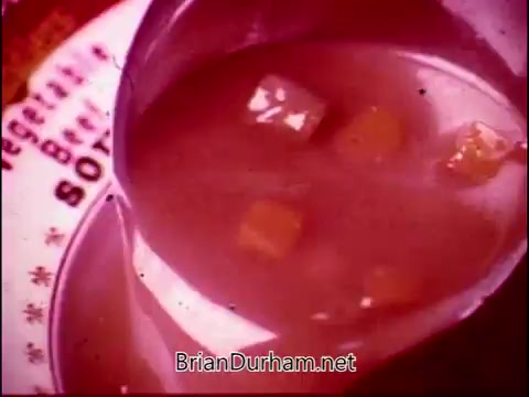 The 16mm still from 'Campbell's Soup - The Manhandlers' features a close-up of a soup being poured from a ladle into a bowl. The soup appears rich and chunky, likely containing vegetables. The image has a warm, vintage color tone, exuding a sense of comfort and nostalgia, while in the background, a plate is partially visible with the words 'Vegetable Beef Soup.' The overall composition highlights the inviting nature of Campbell's Soup, set to the theme song by Frankie Laine.