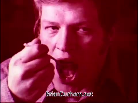 The still from 'Campbell's Soup - The Manhandlers' features a man with short hair, poised to eat from a spoon. The image is tinted with a reddish hue, creating an intense atmosphere. The context highlights a moment of enjoyment, possibly reflecting the themes of camaraderie and comfort associated with food, enhanced by Frankie Laine's theme song.