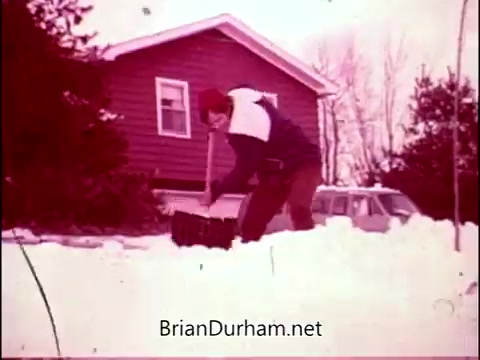 The still depicts a man shoveling snow in front of a modest house, capturing a typical winter scene. The color palette has a faded, nostalgic quality, characteristic of 16mm film. This moment reflects the theme of hard work and resilience, resonating with Frankie Laine's energetic theme song that accompanies 'Campbell's Soup - The Manhandlers.'