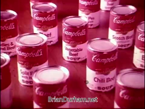 The 16mm still from 'Campbell's Soup - The Manhandlers' features various cans of Campbell's Soup prominently displayed. The cans exhibit familiar branding with bold red labels, showcasing flavors like Vegetable Beef, Chili Beef, and others. The image reflects a nostalgic advertisement atmosphere, highlighting the product with an emphasis on variety and appeal, accompanied by the theme song by Frankie Laine.