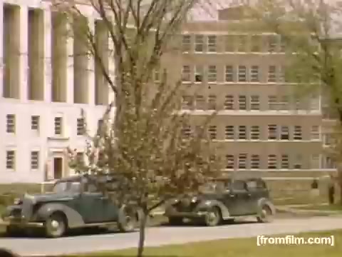 The still from 'Home Movies: Rochester/Niagara Falls 1948-1950' features a street scene with two vintage cars parked along a tree-lined avenue. In the background, notable mid-20th century architecture can be seen, including a large building with columns and a modernist structure. The surrounding trees indicate springtime, highlighting a serene and nostalgic setting from the late 1940s to early 1950s.