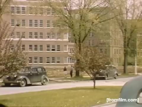 The still from 'Home Movies: Rochester/Niagara Falls 1948-1950' depicts a street scene featuring classic cars from the late 1940s parked along a tree-lined road. In the background, a large brick building with numerous windows suggests an institutional or commercial setting. The surrounding greenery indicates spring or early summer, with blossoming trees adding to the nostalgic atmosphere of the period.