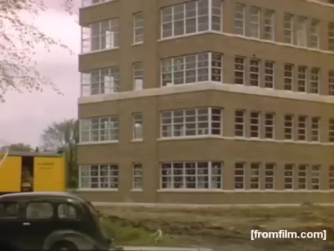 The still from 'Home Movies: Rochester/Niagara Falls 1948-1950' depicts a brick building characterized by large windows, reflecting an architectural style typical of the era. In the foreground, an older model car is parked beside the building, and a yellow vehicle is visible near the entrance, suggesting activity in the area. The scene captures a moment in time, highlighting urban life in mid-20th century America.