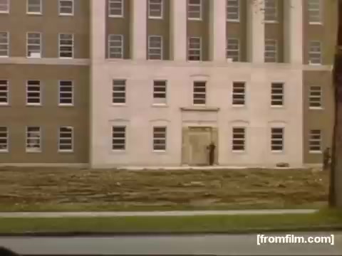 The still from 'Home Movies: Rochester/Niagara Falls 1948-1950' depicts a large, institutional building with a prominent entrance. The structure features a mix of brick and stone, characterized by a classic architectural style with multiple windows. In the foreground, there is an individual standing near the entrance, possibly waiting or observing. The surrounding area appears to have been recently cleared or is under maintenance, suggesting a transitional moment in the landscape or setting. The overall scene reflects a historical period marked by change and development in the region.