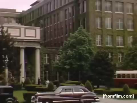 The still shows a historic building, likely a hotel or resort, set in a landscaped area with manicured shrubs and trees. Classic cars from the late 1940s are parked out front, indicating the era. The color palette suggests a warm, nostalgic tone typical of home movies. The scene conveys a sense of leisure and community in mid-20th century Rochester or Niagara Falls.
