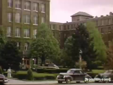 The still from 'Home Movies: Rochester/Niagara Falls 1948-1950' depicts a classic mid-20th century urban scene featuring a large, brick building surrounded by manicured lawns and trees. Vintage cars are parked along the street, contributing to the nostalgic atmosphere of the era. The overall color palette reflects the time period, with a soft, faded quality typical of home movies from the late 1940s to early 1950s.