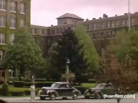 The still from 'Home Movies: Rochester/Niagara Falls 1948-1950' depicts a mid-20th century urban scene featuring multiple trees and greenery in a landscaped area, likely a park or public square. In the foreground, two vintage cars are parked, showcasing the automobile styles of the era. A large building, possibly an institutional or commercial structure, is visible in the background, characterized by its architectural features typical of the period. The overall atmosphere conveys a sense of everyday life in a post-war American city.