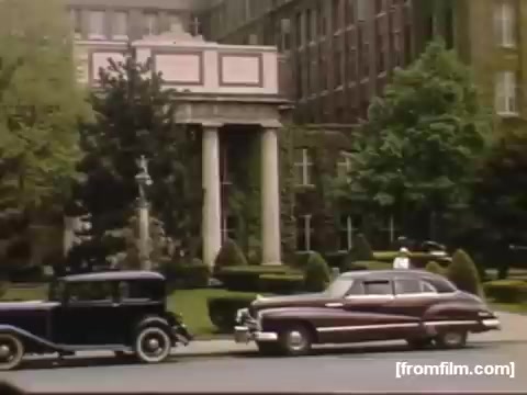 The still captures a scene from the 1948-1950 era, featuring classic cars parked outside a grand building. The architecture exhibits a neoclassical design with notable columns and decorative elements. Lush greenery surrounds the area, enhancing the vintage aesthetic. The image evokes a sense of nostalgia, reflecting everyday life during that time in Rochester/Niagara Falls.
