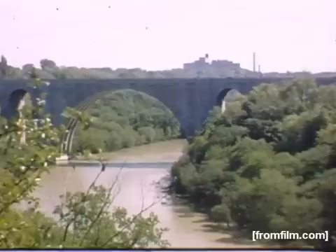 The still captures a scenic view of a bridge arching over a river, surrounded by lush greenery. It's likely set in the Rochester or Niagara Falls area, indicative of the natural beauty and industrial backdrop of the region during 1948-1950. The image conveys a calm, nostalgic atmosphere typical of home movies from that era, highlighting both nature and infrastructure.