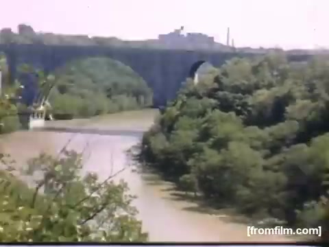 The still depicts a scenic view of a river winding through lush greenery, with an impressive bridge spanning the water. The background features rolling hills and structures, likely indicative of Rochester or Niagara Falls during the late 1940s. The vibrant colors suggest a pleasant season, capturing a snapshot of post-war life and nature in the region.