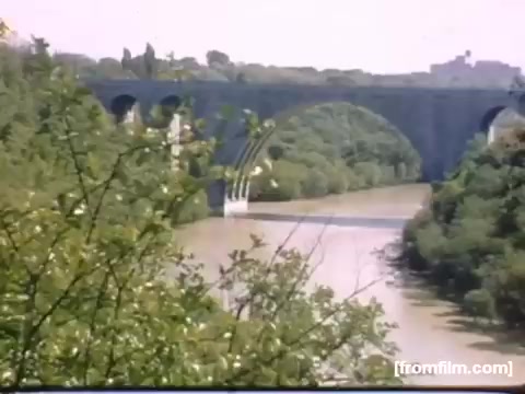 The still depicts a serene landscape featuring a river bordered by lush greenery, with a prominent stone bridge spanning across the water. The scene captures the natural beauty of the area around Rochester and Niagara Falls during 1948-1950, reflecting a tranquil moment in time. The colors are soft and evoke a nostalgic atmosphere typical of home movies from that era.