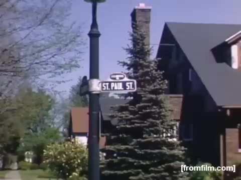 The still features a street sign for 'St. Paul St.' in a residential area, showcasing a brick house and a well-maintained front yard. A large evergreen tree is prominently displayed alongside the sign, suggesting a suburban neighborhood ambiance typical of the late 1940s. The colors are vibrant, characteristic of the film's era, contributing to the nostalgic feel of the home movies.