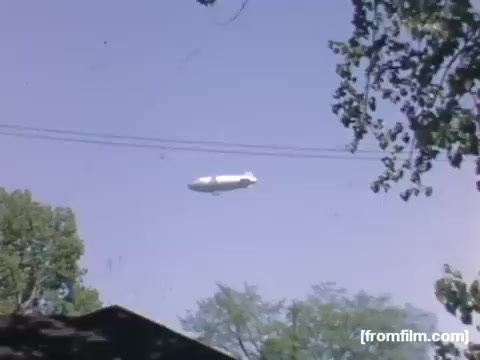 The still from 'Home Movies: Rochester/Niagara Falls 1948-1950' features a blimp floating in a clear blue sky. Surrounding the blimp are green trees, with some outlines of rooftops visible at the bottom. The image captures a moment from the late 1940s, reflecting a leisurely summer day.