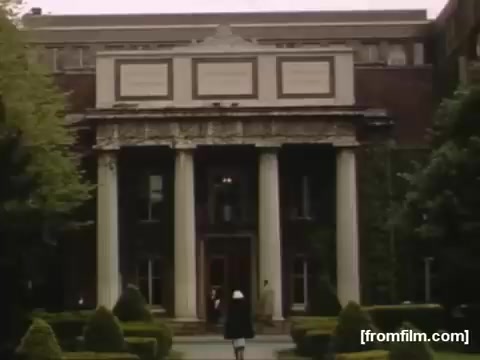 The still from 'Home Movies: Rochester/Niagara Falls 1948-1950' depicts a stately brick building with classical architectural elements, including tall columns and intricate detailing. In the foreground, a figure dressed in attire reminiscent of a nurse or caretaker walks toward the entrance, suggesting a focus on daily life or historical context in the region during that era. The lush greenery surrounding the building enhances its grandeur and provides a serene, welcoming atmosphere.