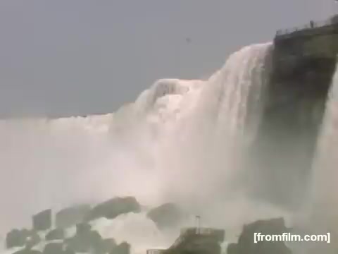 The still features a dramatic view of Niagara Falls, capturing the powerful cascades of water flowing over the edge and creating mist. The rocky foreground suggests the rugged landscape surrounding the iconic site, typical of home movie footage from the late 1940s. The image conveys a sense of natural beauty and dynamism, characteristic of the era's documentary style.