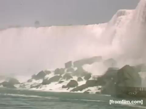 The still captures a scenic view of Niagara Falls, showcasing the powerful cascade of water plunging over the edge. The foreground features rocky formations and mist rising from the falls, highlighting the natural beauty and force of the water. The nostalgic quality of the 16mm film gives the image a vintage feel, evoking the era between 1948-1950 when the footage was taken.