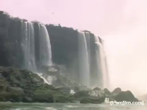 The still depicts the iconic Niagara Falls, showcasing the powerful waterfalls cascading over rocky cliffs. The image captures the natural beauty and dynamic movement of the water, enveloped in a mist that adds an ethereal quality. The colors are soft and slightly faded, reflecting the vintage nature of 16mm film from the late 1940s to early 1950s. This still is part of a nostalgic collection that emphasizes personal experiences and memories associated with the region.