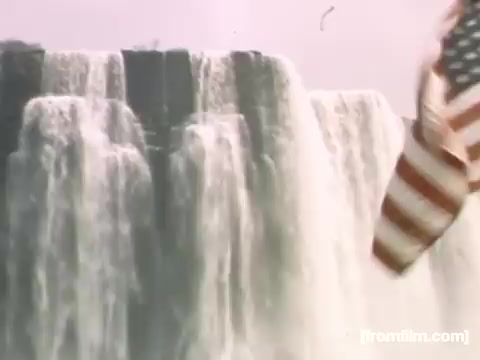 The still captures a scene from 'Home Movies: Rochester/Niagara Falls 1948-1950,' showcasing the majestic Niagara Falls. Water cascades dramatically over the edge, while an American flag waves in the foreground, symbolizing a connection to the location and its cultural significance. The image reflects the natural beauty and historical context of the era.