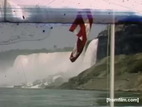 The still captures a scene from the documentary 'Home Movies: Rochester/Niagara Falls 1948-1950.' It features a view of Niagara Falls, with the iconic waterfalls in the background and an American flag visible in the foreground. The image has a vintage quality, reflecting the 16mm film format, and shows subtle imperfections typical of home movies from that era, such as scratches and faded colors. The setting evokes a nostalgic atmosphere, highlighting both the natural beauty of the falls and a moment of personal memory.