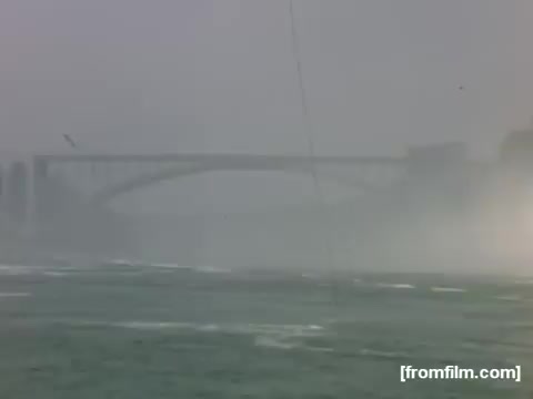The still from 'Home Movies: Rochester/Niagara Falls 1948-1950' captures a misty scene of Niagara Falls, with the iconic bridge partially obscured by fog and spray. The atmosphere is serene yet dynamic, highlighting the natural beauty and power of the falls during that era. The muted colors contribute to a nostalgic feel, evoking the historical context of post-war America.