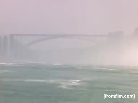 The 16mm still from 'Home Movies: Rochester/Niagara Falls 1948-1950' depicts a misty scene near Niagara Falls. In the foreground, the turbulent waters are visible, with whitecaps indicating strong currents. A bridge arches in the background, partially obscured by fog, conveying a sense of natural power and the iconic landmark's atmosphere. The color palette suggests a soft, nostalgic view characteristic of mid-century home movies.
