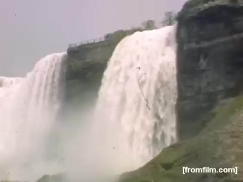 The still depicts a scenic view of Niagara Falls, showcasing the powerful flow of water cascading over the cliffs. The lush greenery at the base contrasts with the mist rising from the falls, emphasizing the natural beauty of the area. The image captures a moment of the mid-20th century, reflecting the iconic tourist destination during the late 1940s.