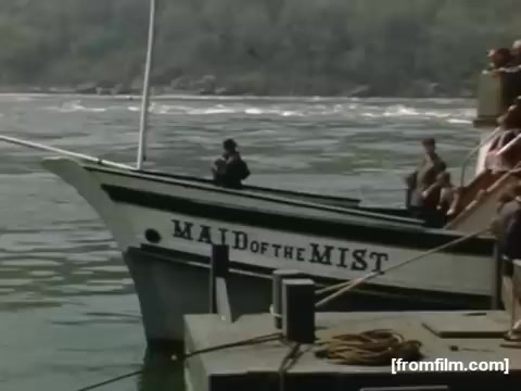 The still features the 'Maid of the Mist,' a boat situated at the dock near Niagara Falls. It shows passengers, some wearing hats and jackets, preparing to board. The backdrop includes the river and lush greenery, reflecting the mid-20th-century atmosphere of the area. The image captures a moment of leisure and adventure typical of this iconic tourist experience.
