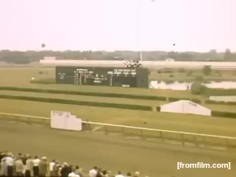The still from 'Home Movies: Rochester/Niagara Falls 1948-1950' captures a scene at a horse racing track. In the foreground, a crowd watches the race, while a scoreboard displays race information in the background. The landscape features green grass, a clear sky, and various structures related to the racetrack, conveying a lively atmosphere typical of mid-20th century American leisure activities.