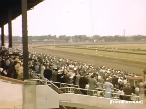 The still from 'Home Movies: Rochester/Niagara Falls 1948-1950' captures a lively scene at a horse racing track. A large crowd is gathered in the grandstand, watching the races. The atmosphere is vibrant, with a mix of fashion typical of the late 1940s. The racetrack extends into the distance, suggesting excitement and anticipation as horses compete. The image conveys a sense of community and leisure from that era.