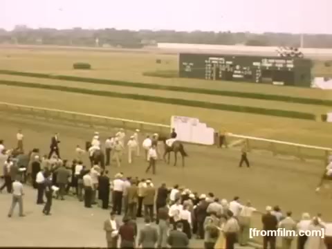 The still captures a bustling horse racing scene from the late 1940s. A crowd gathers around the racetrack, watching as jockeys and their horses prepare for a race. In the background, a scoreboard displays race information, and the atmosphere conveys excitement and anticipation typical of the era's social events. The colors and fashion of the attendees reflect the style of the time.