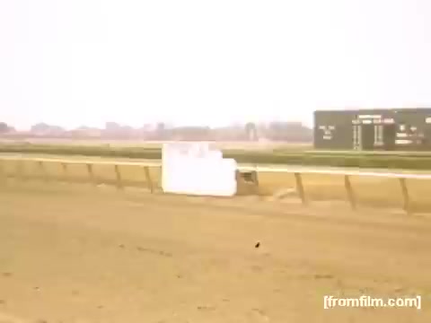 The still from 'Home Movies: Rochester/Niagara Falls 1948-1950' depicts a scene at a horse racing track. The foreground shows a sandy racetrack, while a blank white sign is positioned near the track's edge. In the background, a scoreboard displays race information, indicative of the vibrant racing culture of the time. The overall atmosphere captures a nostalgic, everyday moment from mid-20th century American life.