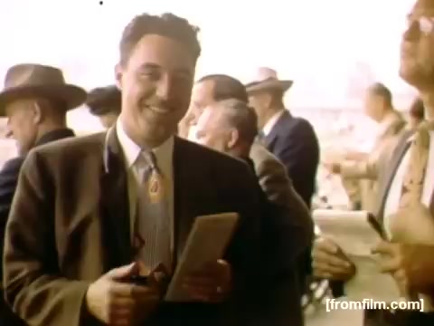 The still depicts a smiling man in 1940s attire, holding a notepad, surrounded by other men in similar clothing, likely at an outdoor event. The setting suggests a social or public gathering, possibly related to leisure activities or a competition, with a nostalgic color palette characteristic of the era.