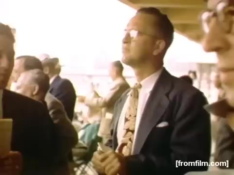 The still from 'Home Movies: Rochester/Niagara Falls 1948-1950' depicts a group of men, likely in a social or public setting. One man in the foreground stands out, wearing glasses and a stylish suit with a patterned tie, looking contemplatively upwards. The atmosphere appears to be relaxed and sociable, typical of mid-20th century leisure moments. The color palette reflects the vintage quality of the film, emphasizing a nostalgic perspective of the period.