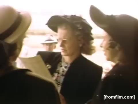 The still from 'Home Movies: Rochester/Niagara Falls 1948-1950' depicts three women, likely engaged in conversation. One is holding a document or piece of paper, while the others are attentively listening. The scene conveys a sense of leisure and social interaction typical of the era, with stylish hats indicating a time of fashionable outings. The background hints at a summer day, contributing to the nostalgic feel of the home movie genre.