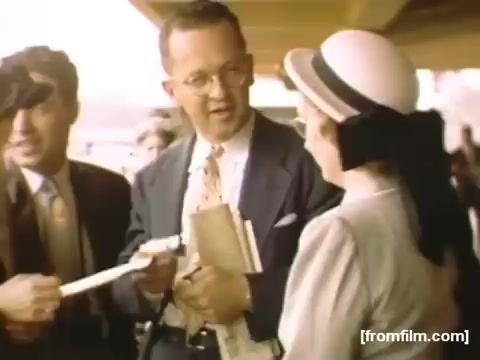 The still from 'Home Movies: Rochester/Niagara Falls 1948-1950' captures a moment at a transportation hub, likely an airport or train station. A man in a suit, holding papers, is engaging with a woman dressed in a vintage outfit, complete with a wide-brimmed hat. A second man stands nearby, appearing to observe the interaction. The scene conveys a nostalgic atmosphere, reflecting social interactions of the late 1940s through its clothing and setting.