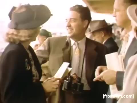 The still from 'Home Movies: Rochester/Niagara Falls 1948-1950' depicts a group of people in a social setting. A well-dressed man is smiling and interacting with a woman who is wearing a stylish hat and dressed in black. He holds a camera around his neck, suggesting an interest in capturing moments. The background features other individuals in similar attire, hinting at a lively gathering during that era. The overall atmosphere conveys a sense of nostalgia and connection among the group.