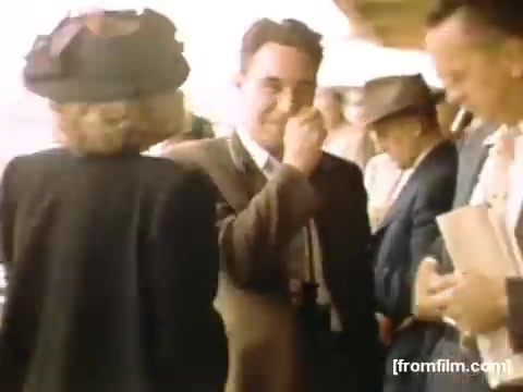 The still from 'Home Movies: Rochester/Niagara Falls 1948-1950' shows a group of people in a casual outdoor setting. A man in a suit smiles while interacting with a woman in a hat and black attire. Other individuals are visible in the background, engaged in conversation. The scene captures a moment of social interaction and reflects the fashion and communal ambiance of the late 1940s. The color tones suggest a nostalgic and warm atmosphere typical of home movies from that era.