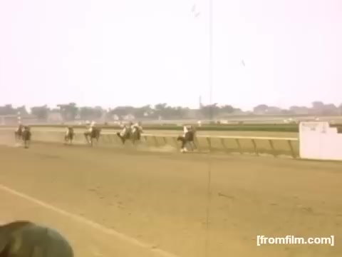 The still captures a lively horse race taking place on a dirt track. Several jockeys can be seen riding their horses, vying for position while spectators watch in the background. The scene reflects the mid-20th century atmosphere, typical of the time when the film was made, showcasing a moment of excitement and competition in Rochester or Niagara Falls. The soft colors and slightly faded quality evoke a nostalgic feel of home movies from that era.