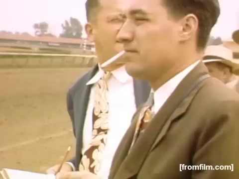 The still captures two men at a racetrack in the late 1940s, likely observing the events unfolding. One man is dressed in a suit and holds a notepad, suggesting he might be jotting down notes or placing bets. The other man, slightly behind, smokes a cigarette. The color tint adds a nostalgic feel, characteristic of home movies from the era. The background features a racetrack, hinting at the leisure activities of that time.