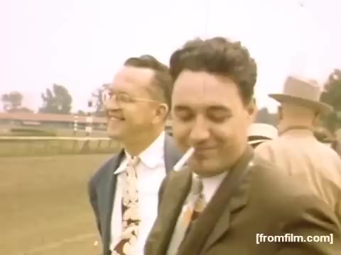 The still captures two men standing outdoors, likely at a racetrack, smiling and enjoying the moment. One man wears glasses and has a broad smile, while the other, dressed in a suit, has a cigarette in his mouth and a relaxed expression. The background shows a gathering of people and a blurred outline of a venue, suggesting a lively social atmosphere typical of the late 1940s. The color palette has a warm, vintage tone characteristic of 16mm film from that era.