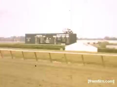 The still captures a scene from a racetrack, likely during a horse racing event. A large scoreboard displays race information, with a dirt track in the foreground. The atmosphere is tranquil, indicative of the era between 1948-1950 in Rochester/Niagara Falls, suggesting a leisurely day at the races. The colors appear muted, typical of vintage film.