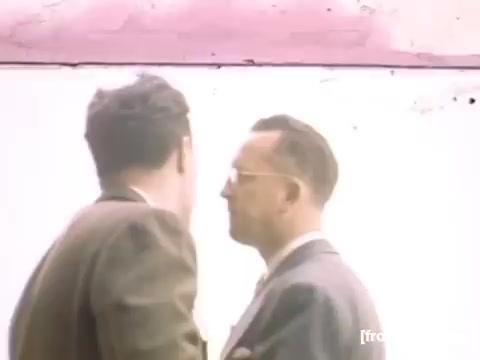 The still from 'Home Movies: Rochester/Niagara Falls 1948-1950' captures two men engaged in a close conversation against a pale background. The scene likely reflects intimate social interactions typical of the era, showcasing their attire—formal suits indicative of the period. The muted colors and soft focus evoke a nostalgic ambiance characteristic of home movie footage from the late 1940s.