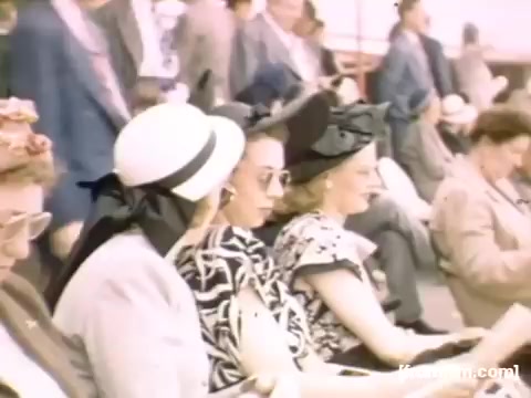 The still from 'Home Movies: Rochester/Niagara Falls 1948-1950' features a group of elegantly dressed women seated in a public setting. They are wearing fashionable hats and sunglasses, indicative of the era's style. The background is filled with other attendees, creating a lively atmosphere, likely at a social event or gathering during that time. The color and presentation reflect the vintage aesthetic typical of mid-20th century home movies.