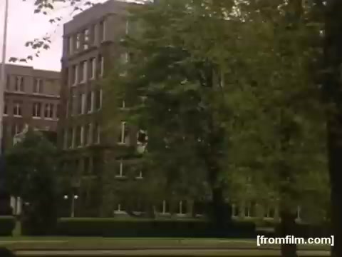 The still from 'Home Movies: Rochester/Niagara Falls 1948-1950' captures a view of a brick building set amidst lush greenery. The architecture appears mid-20th century, with large windows and a formal landscaping style, indicative of the period. The scene suggests a serene urban environment typical of the Rochester or Niagara Falls area during that era.