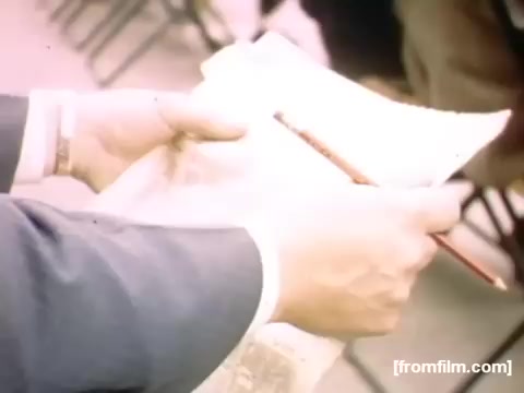 The still features a close-up of a person's hands holding a newspaper and a pencil. The newspaper appears to be folded and the scene conveys a sense of everyday life in Rochester or Niagara Falls during the late 1940s. The composition suggests engagement with current events or personal notes, capturing a candid moment from the era. The warm color tones enhance the nostalgic feel of the 16mm film.