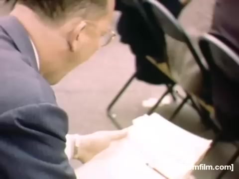 The still depicts a man in a suit, intently looking down at a document or notebook while seated in a gathering, likely during a meeting or event. His focus suggests he is engaged with the material, indicative of the social or professional context of the time. The background features other seated individuals, hinting at a group setting. This scene captures a moment of everyday life in the late 1940s, reflecting the culture and formalities of the era.