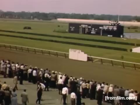 The still depicts a lively scene at a horse racing event, showcasing a large crowd gathered along the track. Spectators, dressed in mid-20th-century attire, are focused on the race, with a scoreboard in the background displaying race information. The image captures the vibrant atmosphere of post-war America during the late 1940s in Rochester/Niagara Falls.